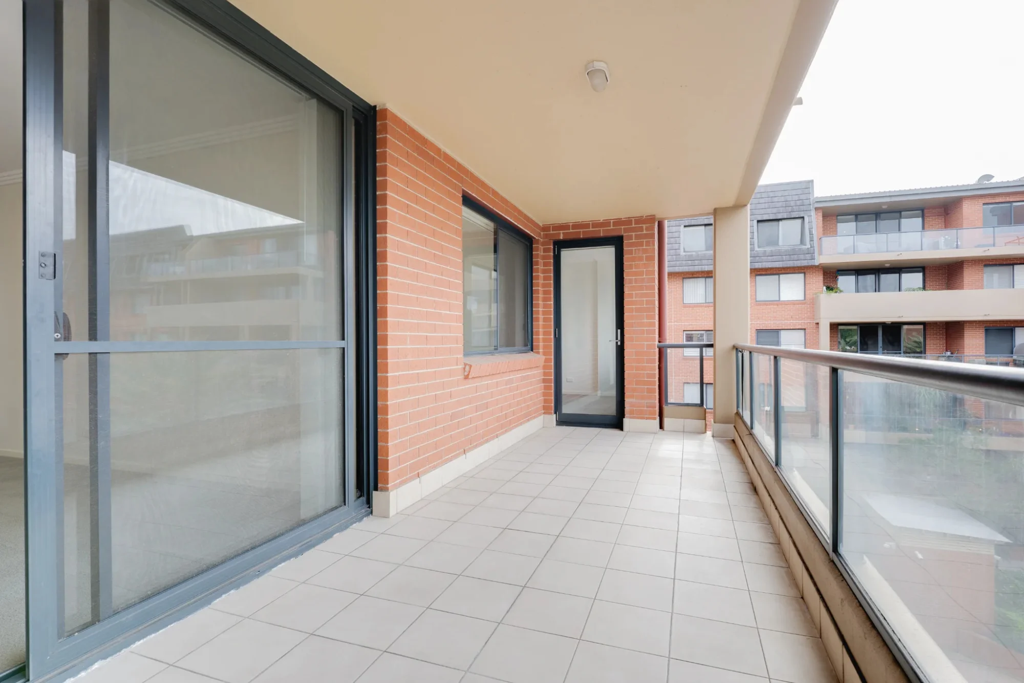 An empty apartment balcony before staging: bare tile, a glass railing, and a view of the building opposite.