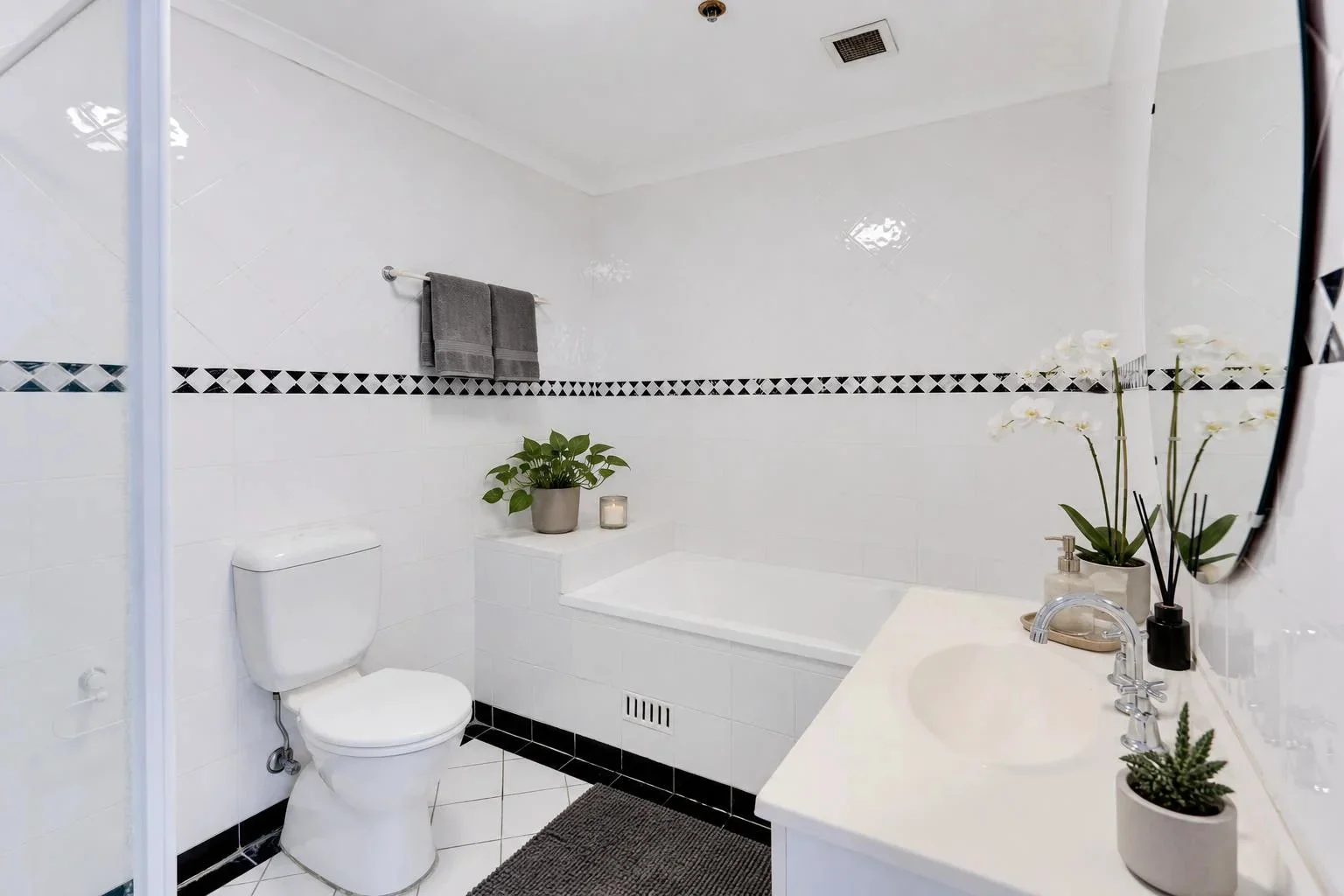 The same bathroom staged with SecondLight: towels, an orchid, a candle, and a bath mat added, with the fixtures and mirror reflection left untouched.